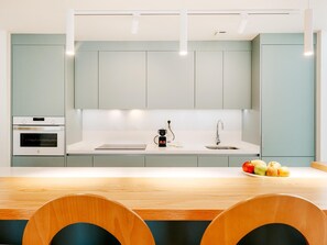 A modern kitchen with light green cabinets, a white countertop, a built-in oven, a coffee maker, and a sink. There are two wooden chairs at the table.