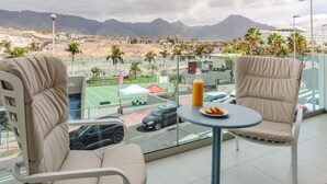 A balcony with two beige cushioned chairs and a small round table with a bottle and a plate of food. A tennis court is visible in the background.