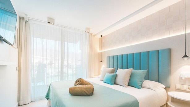 A modern hotel room with a large bed, a bedside table, and a view through sheer curtains.