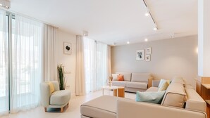 A modern living room with a sofa, armchair, and ottoman, large windows with sheer curtains, and a minimalist decor.