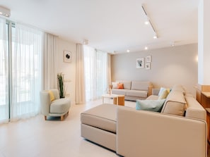 Uma sala de estar moderna com sofá, poltrona e banquinho, janelas amplas com cortinas leves e decoração minimalista.