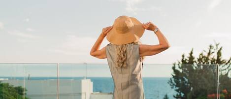 A person standing on a balcony overlooking a body of water, wearing a hat and a sleeveless dress.