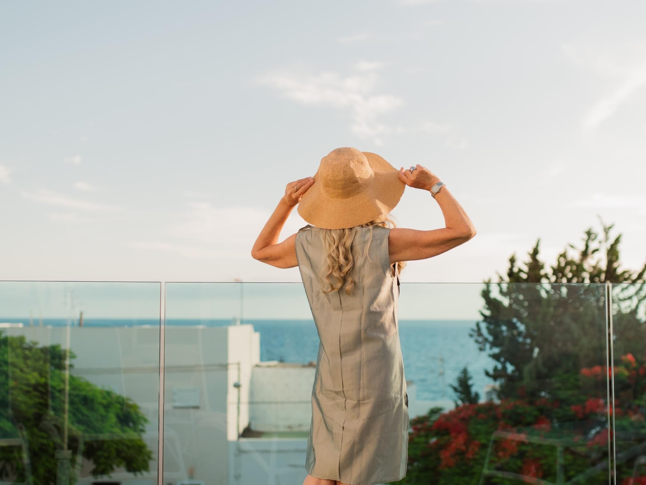 A person standing on a balcony overlooking a body of water, wearing a hat and a sleeveless dress.