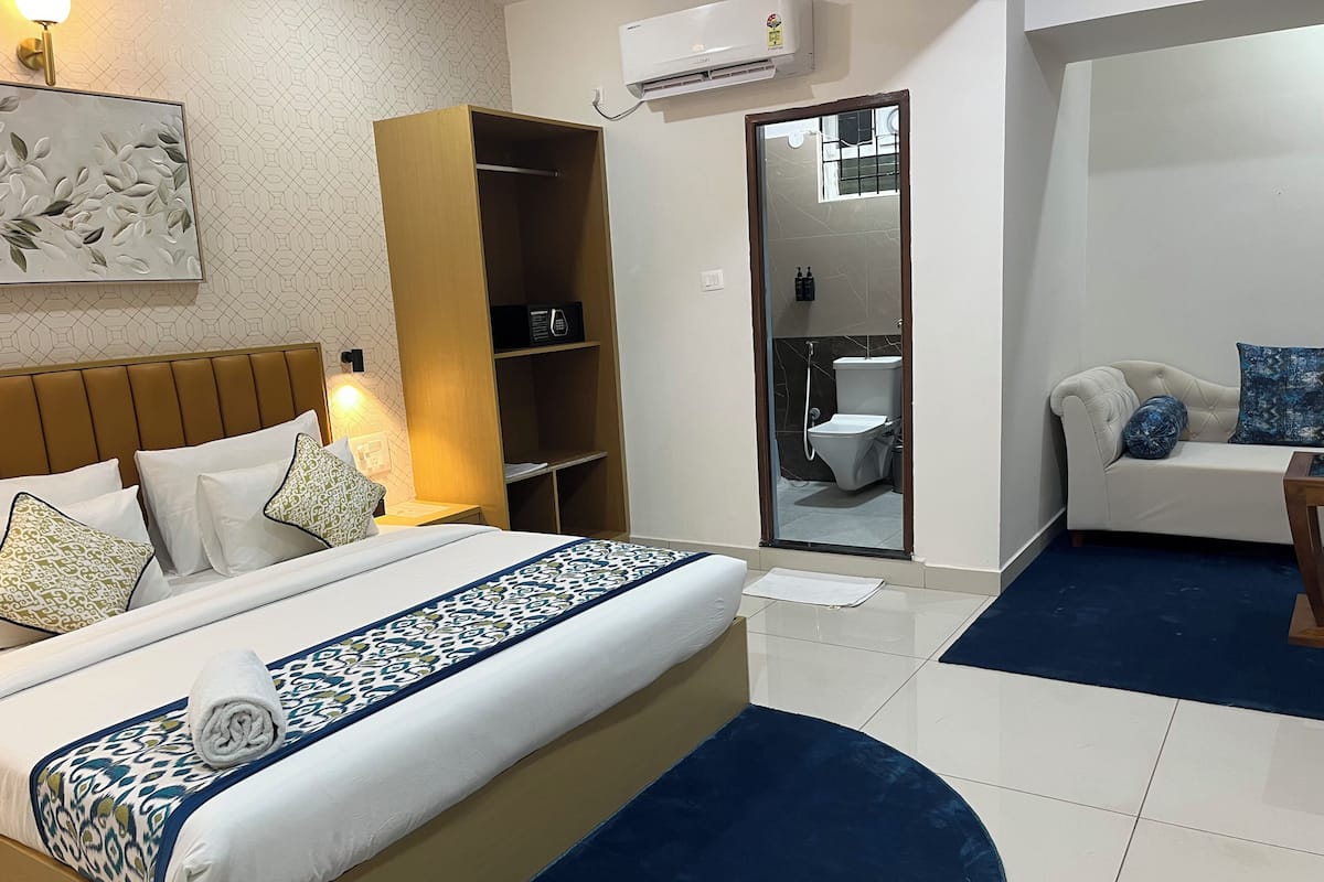 A hotel room with a bed, a wardrobe, a sofa, and a bathroom visible through a glass door.