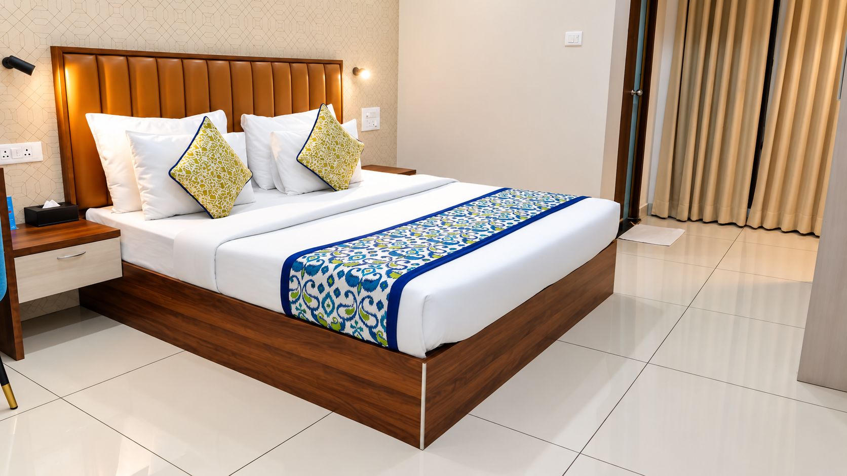 A modern hotel room with a wooden headboard, a neatly made bed with patterned bedding, a bedside table with a lamp, and a view of the outside through a window with curtains.