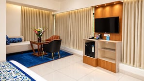 Quarto de hotel moderno com sofá, mesa de jantar com cadeiras, pequena geladeira, televisão de tela plana e tapete azul.