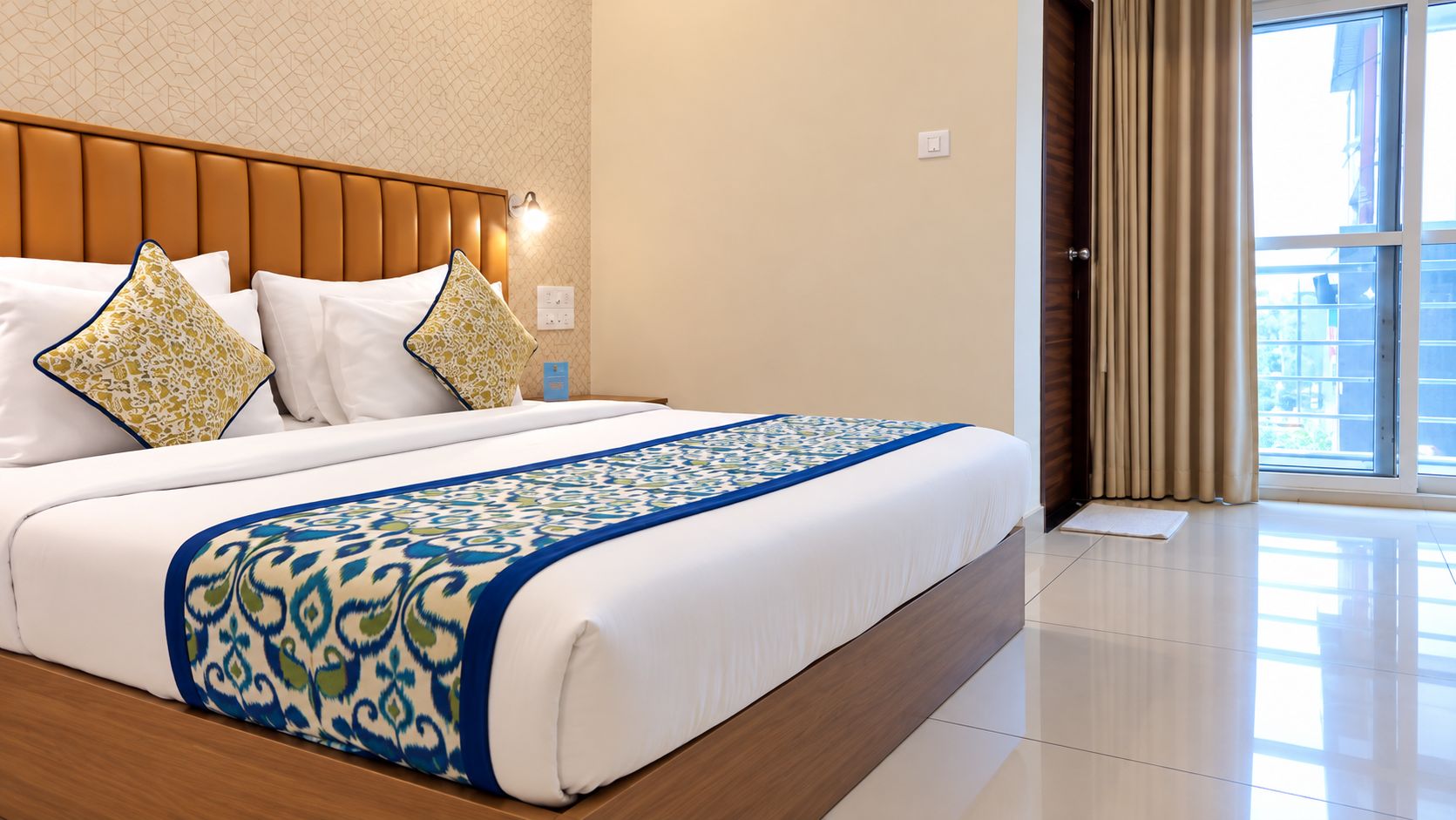 A hotel room with a large bed, a wooden headboard, a patterned bedspread, and a view of the outside through a window with curtains.