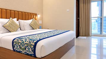Quarto de hotel com uma cama grande, cabeceira de madeira, colcha estampada e vista para o exterior através de uma janela com cortinas.