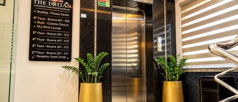 An elevator with a sign listing hotel floors and room numbers, a reception area, and two potted plants.