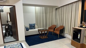 Habitación de hotel moderna con sofá, dos sillones, una mesita, un minibar y vistas a la calle a través de cortinas.