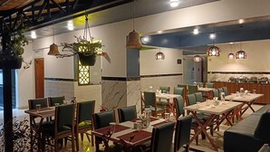 Ein Restaurant mit mehreren Tischen, Stühlen, hängenden Pendelleuchten und einer sichtbaren Küchenzeile im Hintergrund.