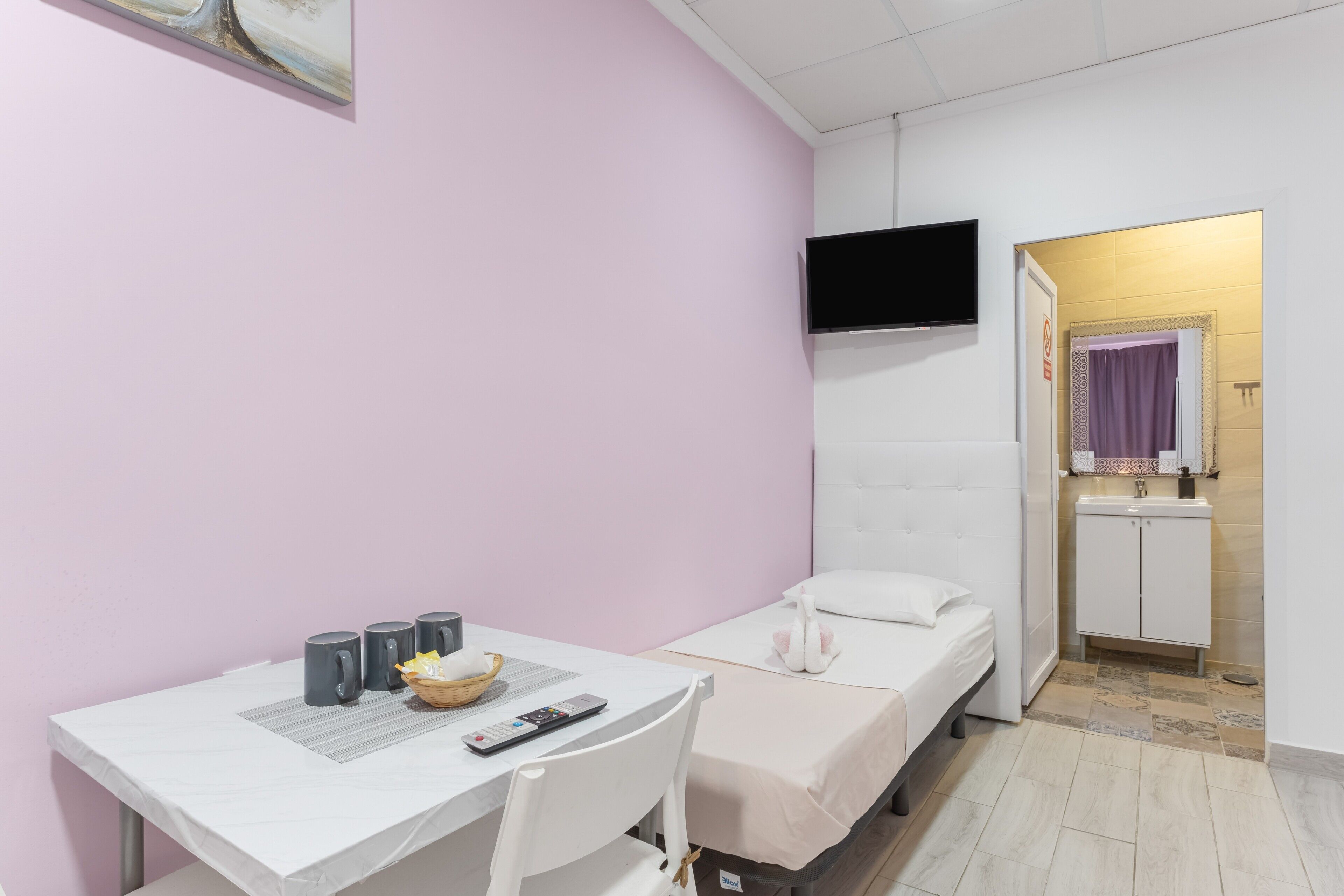 A small room with a bed, a desk with a chair, a television mounted on the wall, and a bathroom visible through an open door.