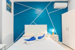 Quarto de hotel com parede azul geométrica, cama branca, mesa de cabeceira com abajur e guarda-roupa.