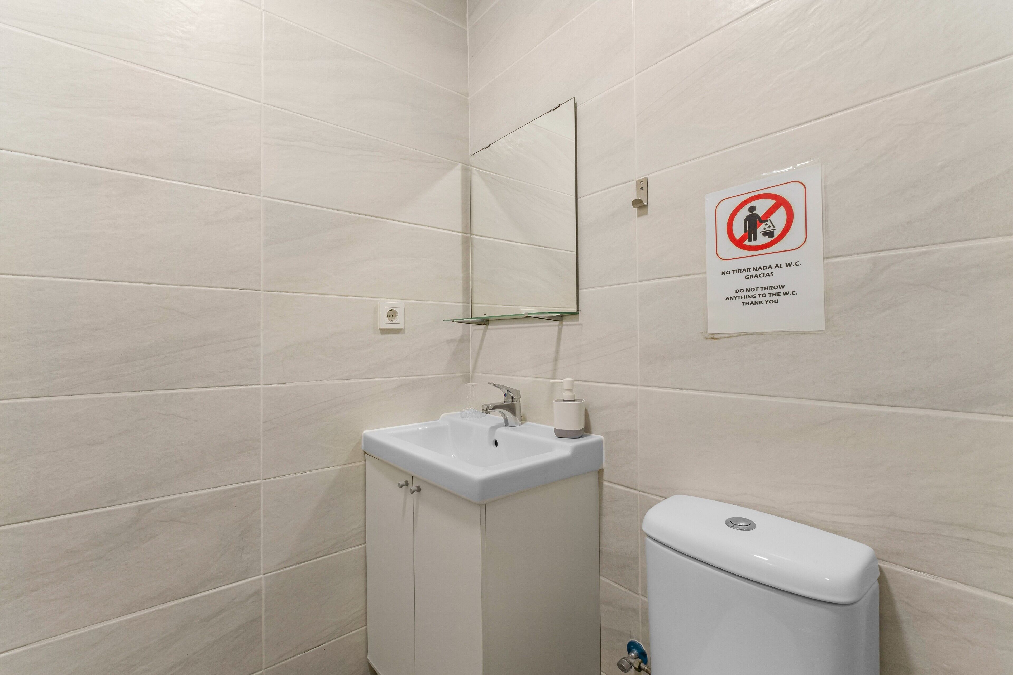 Et badeværelse med en hvid vask, spejl og toilet, samt et skilt, der forbyder bortskaffelse af affald.