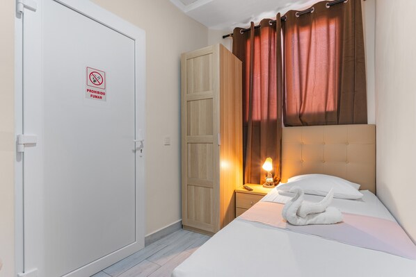 A hotel room with a bed, a nightstand with a lamp, a wooden wardrobe, and a door with a 'No Smoking' sign.