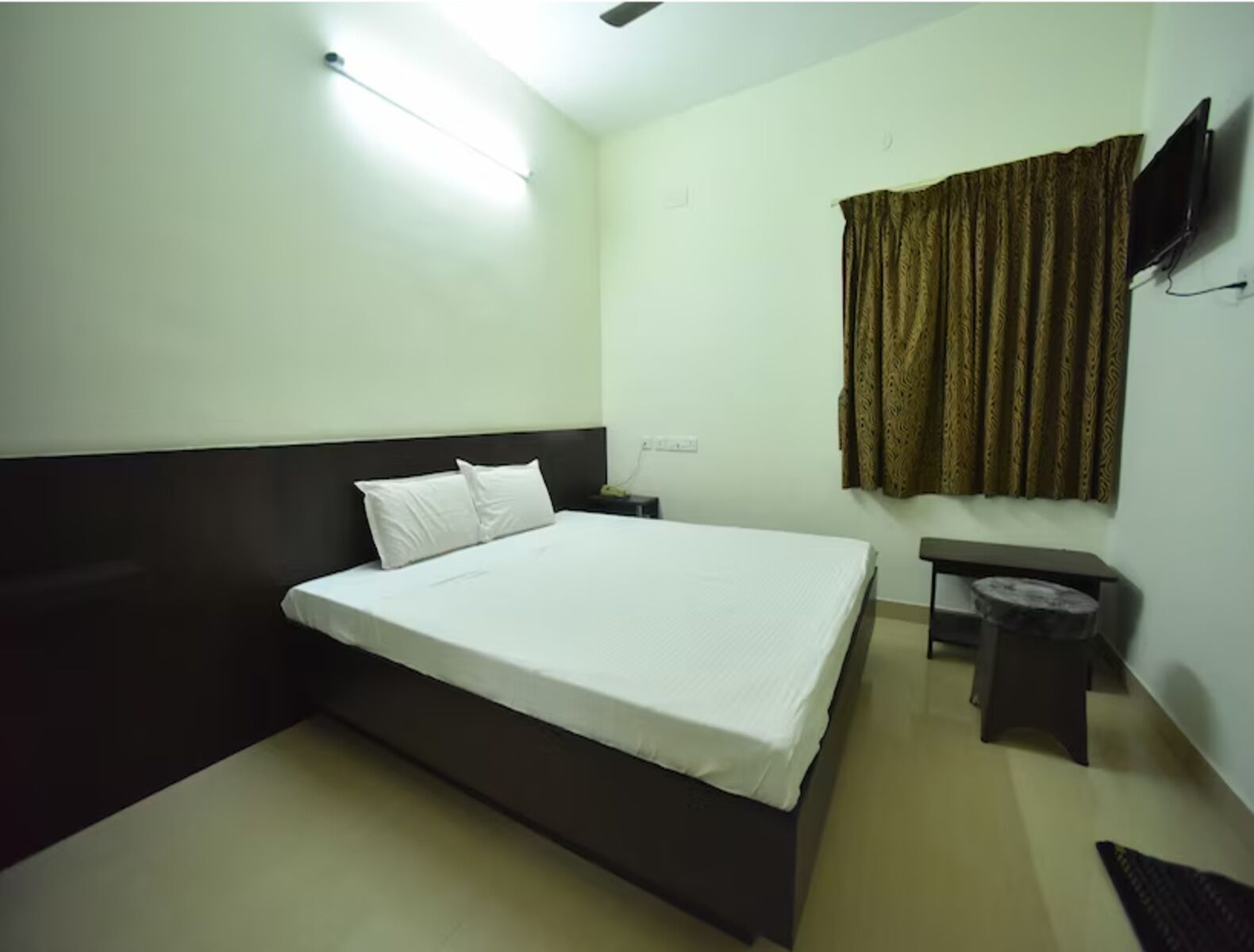 Habitación de hotel con una cama individual, una mesita de noche, un televisor montado en la pared y una ventana con cortinas.