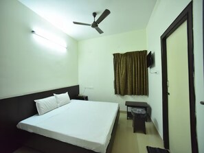 Habitación de hotel con una cama individual, ventilador de techo, televisor montado en la pared y una pequeña mesa con una silla.