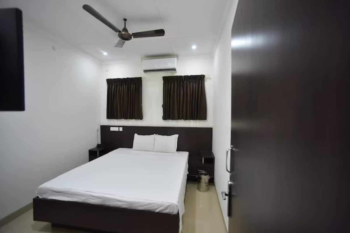 Una habitación de hotel con una cama, un ventilador de techo, un equipo de aire acondicionado, una mesita de noche, una puerta y un televisor fijado a la pared.