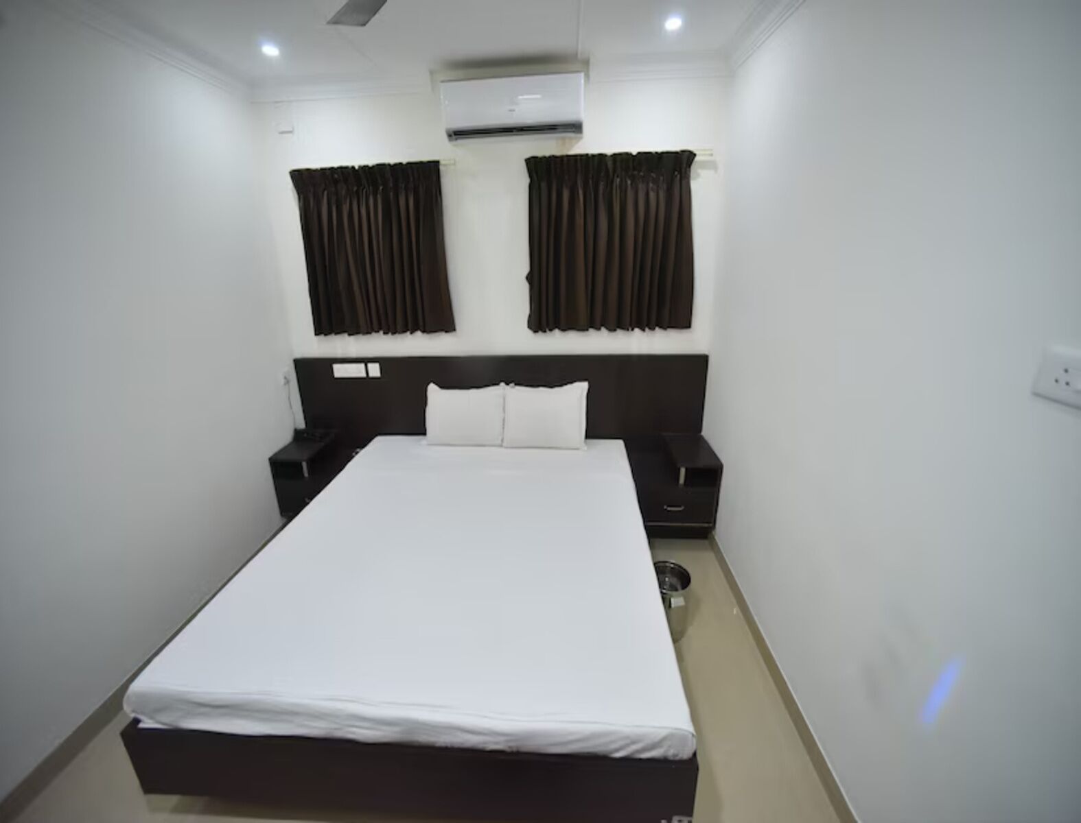 Una habitación de hotel con una cama grande, dos mesitas de noche, un aire acondicionado y cortinas oscuras.