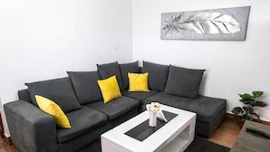 Un salon moderne avec un canapé d’angle gris, des coussins jaunes, une table basse blanche et un tableau abstrait gris accroché au mur.