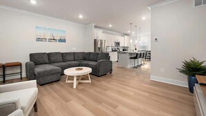 A modern living room with a grey sectional sofa, a round coffee table, and a white armchair. A kitchen with white cabinets and stainless steel appliances is visible in the background. - Z Estates | Balcony Beach View Townhome + Pool (Ocean City)