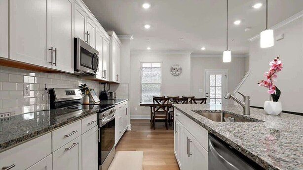 A modern kitchen with white cabinets, granite countertops, a microwave, and a dining area with a wooden table and chairs. - Z Estates | Balcony Beach View Townhome + Pool (Ocean City)