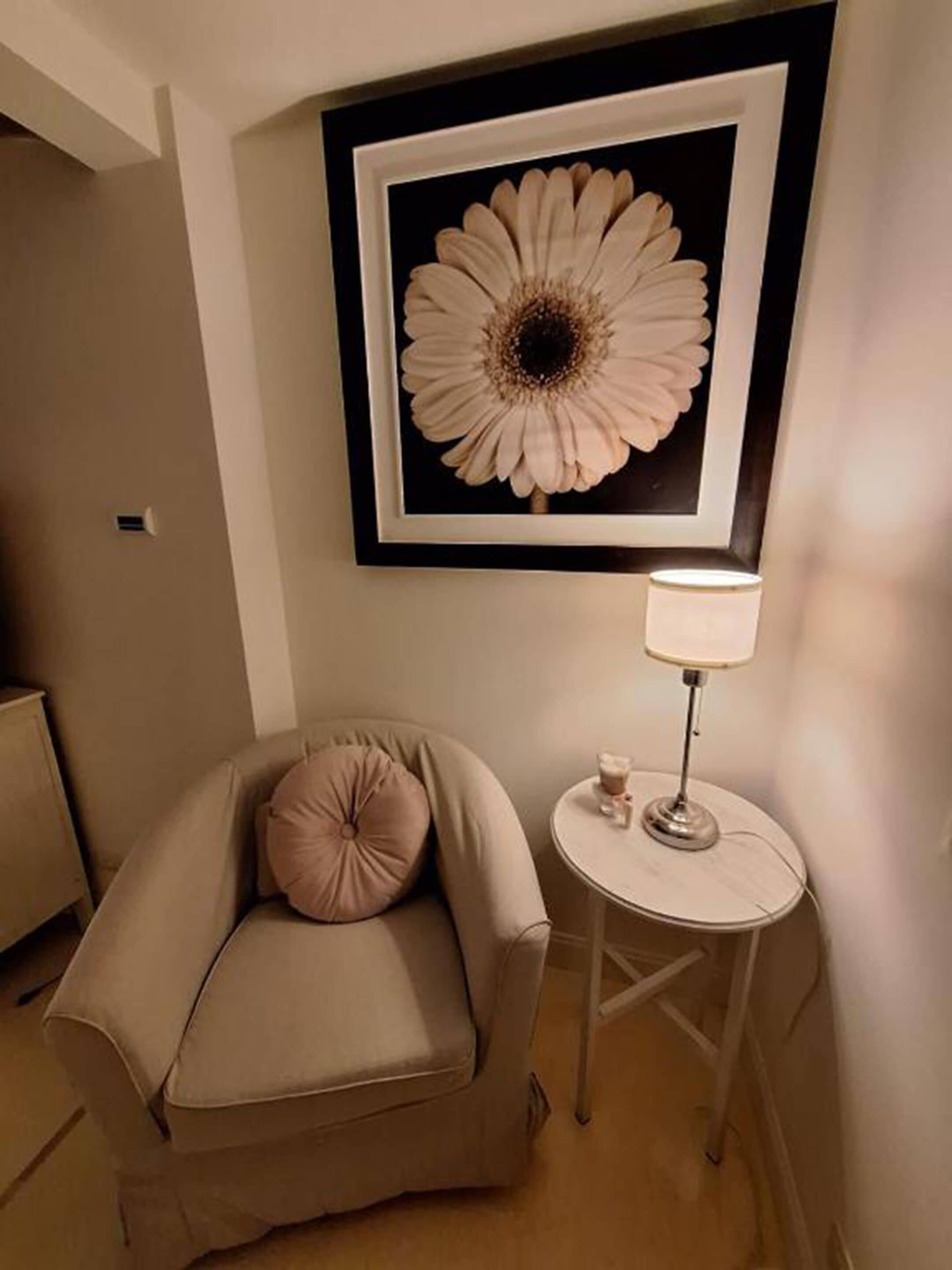 Een kamer met een beige fauteuil, een ronde tafel met een lamp en een bloemkunstwerk in een lijst aan de muur.