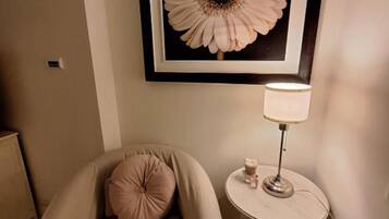Een kamer met een beige fauteuil, een ronde tafel met een lamp en een bloemkunstwerk in een lijst aan de muur.