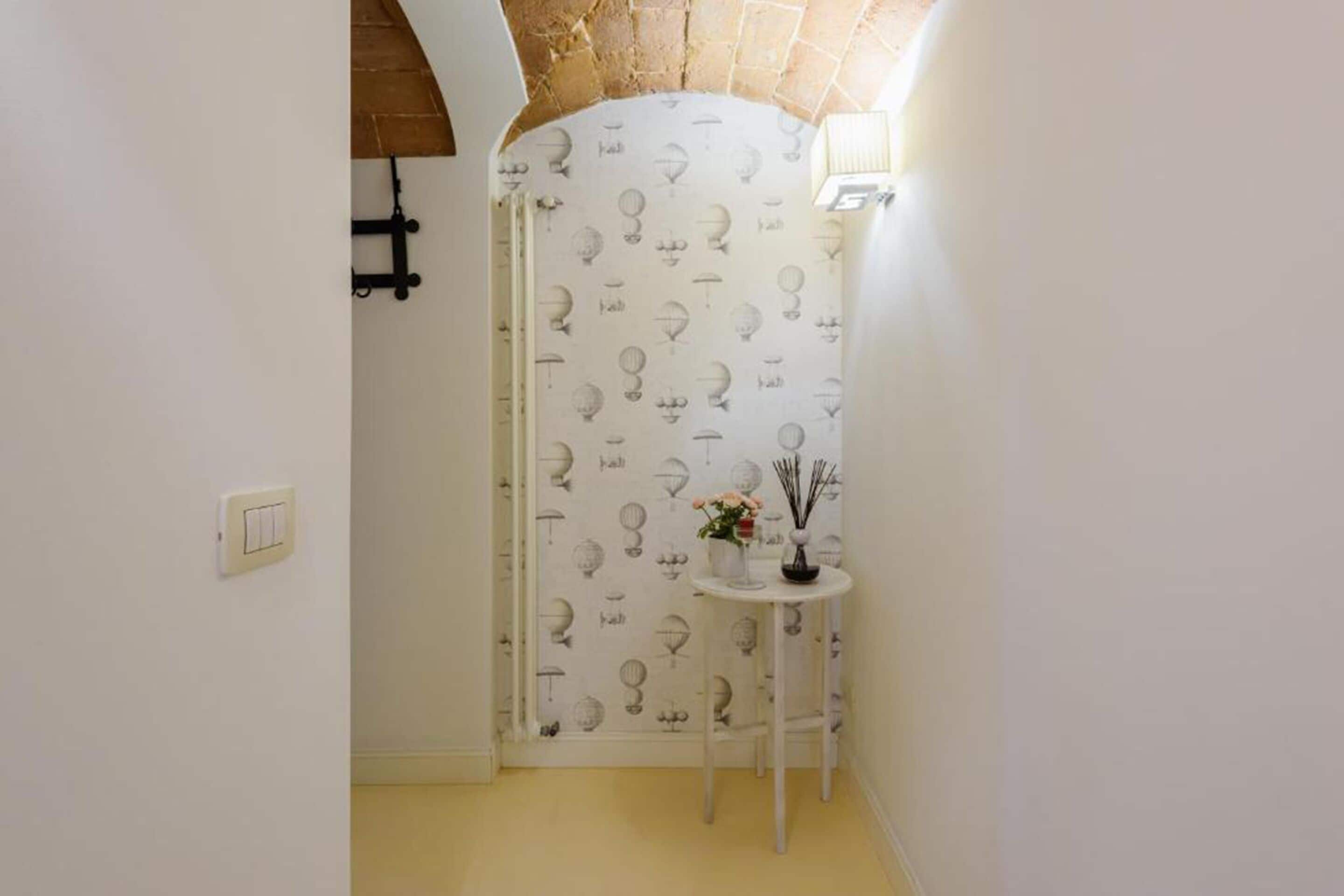 Ein schmaler Flur mit einer weißen Tür, verziert mit einem Muster aus Glühbirnen. An der Wand steht ein kleiner weißer Tisch mit einem Blumenstrauß und einer Kerze.
