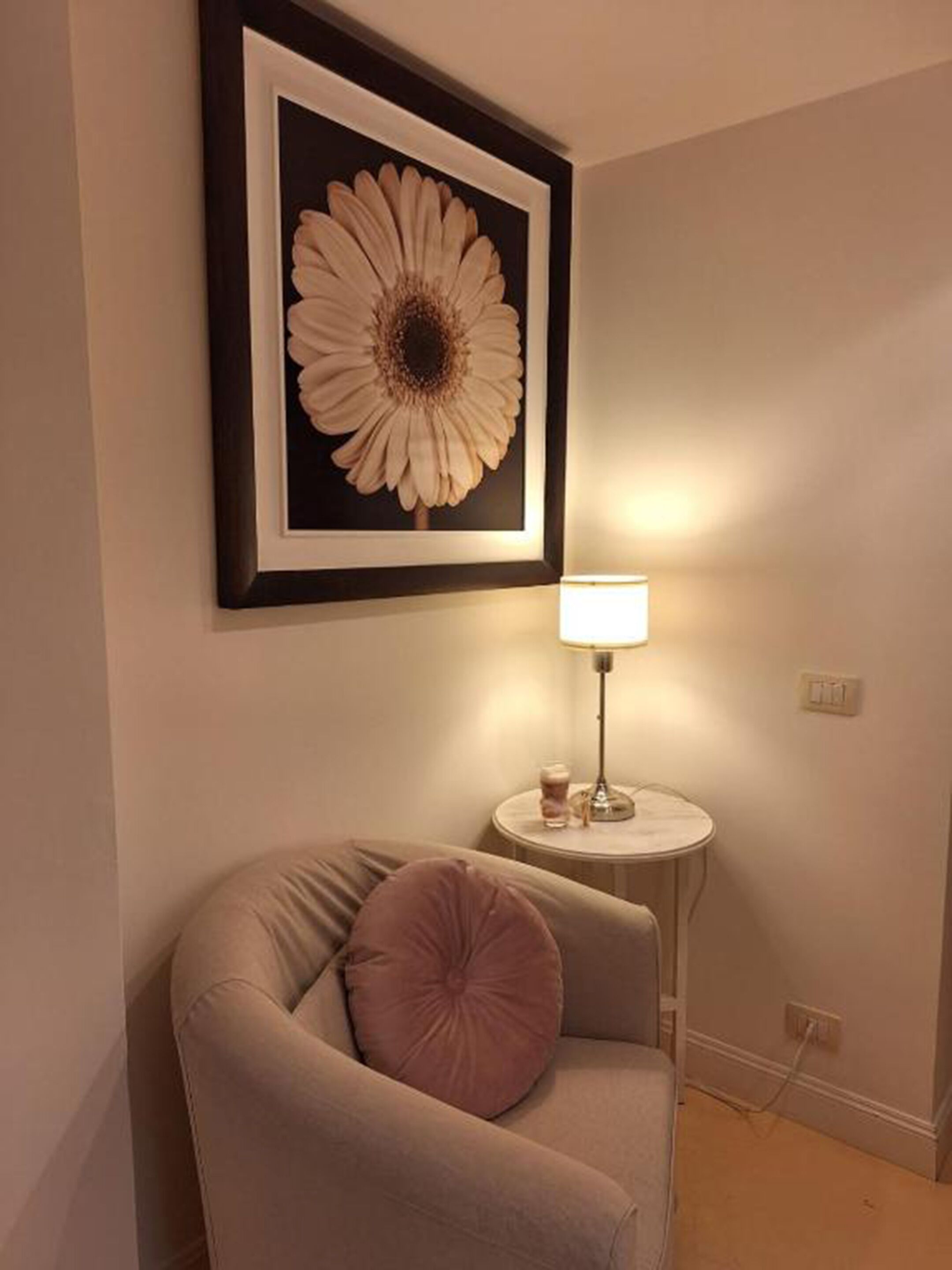 A corner room with a beige armchair, a round side table with a lamp, and a framed flower artwork on the wall.