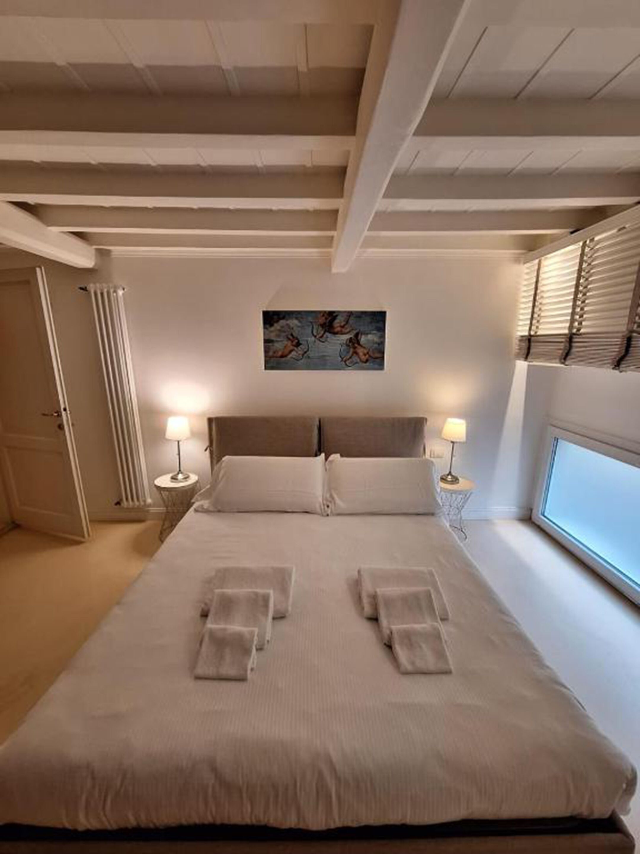 A bedroom with a large bed, two bedside lamps, a radiator, and a window with blinds.
