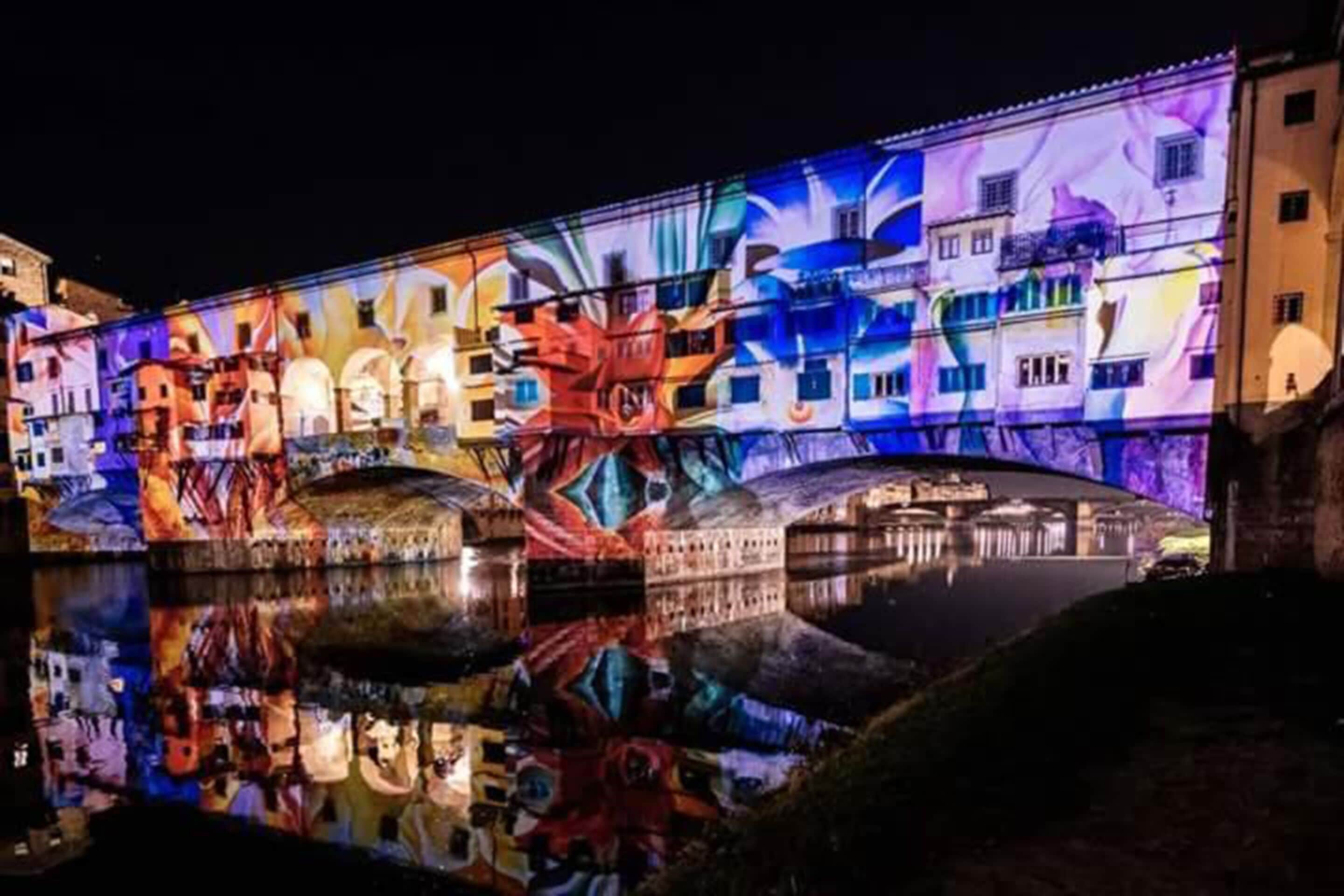 Un edificio con un mural colorido en la fachada, un puente en primer plano y una masa de agua que refleja la escena.