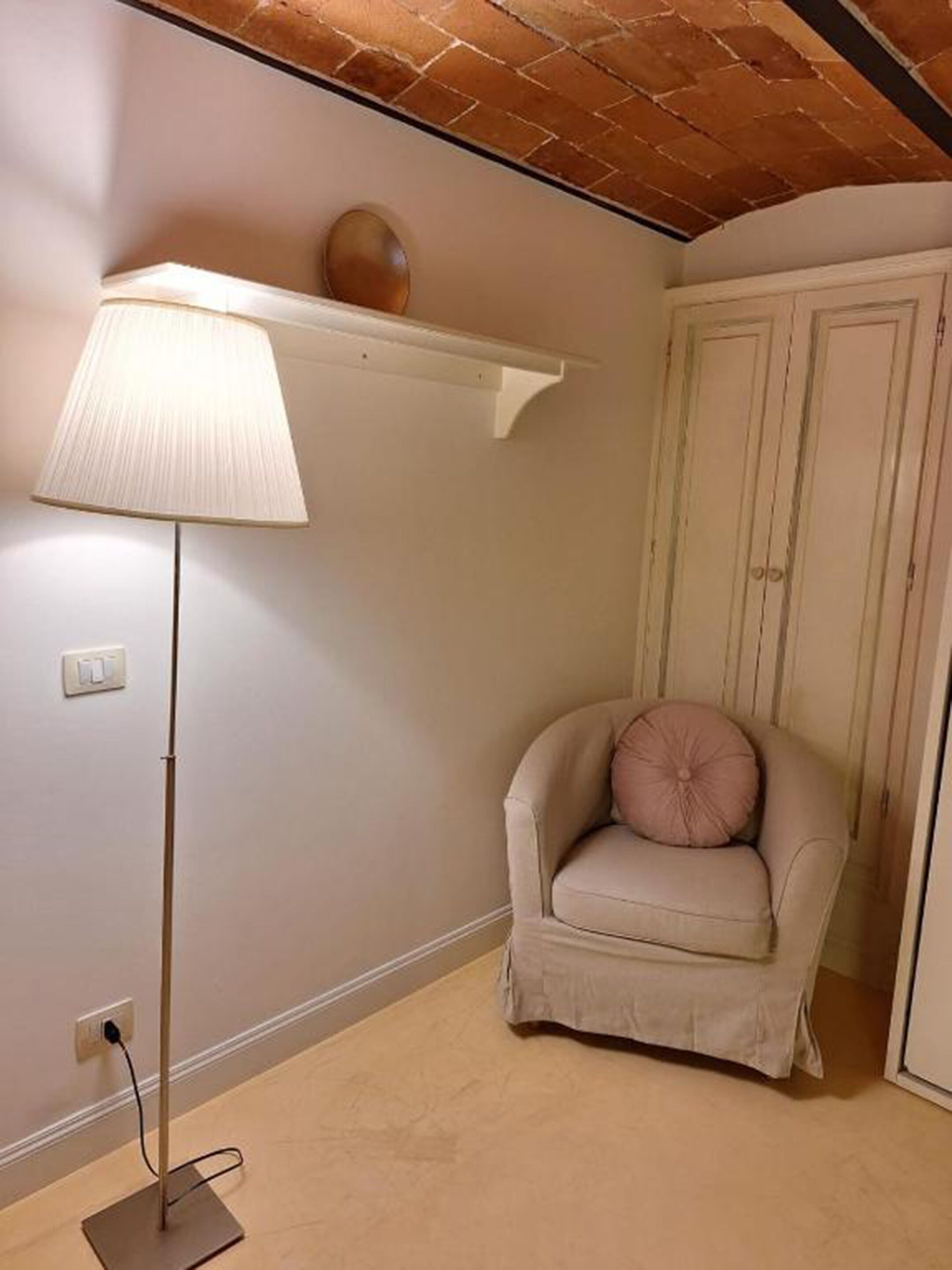 A room with a beige floor, a standing floor lamp, a beige armchair with a cushion, a white door, and a brick ceiling.