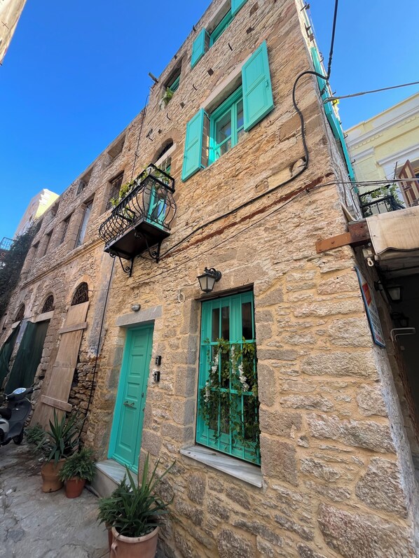 A stone building with green shutters, a balcony with a wrought iron railing, and potted plants on the ground floor.