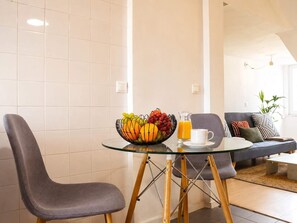 A modern dining area with a glass table, two grey chairs, a fruit bowl, a glass pitcher of orange juice, and a white mug.