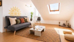 A modern loft apartment with a sloped ceiling, a grey sofa, a coffee table, a potted plant, and a small window. - Sunset Terrace • Tagus Views • Walk to LX Factory (Lisbon)