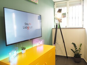 A room with a TV displaying 'StayIN Lisbon.pt', a yellow desk with a neon 'hello' sign, a potted plant, and a window with blinds. - Ground-Floor 1BR Merces• Chiado •  Tram 28 (Lisboa)