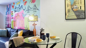 Een gezellige zithoek met een bank, een glazen tafel met eten en drinken, en levendige kunstwerken die Lissabon en een straatbeeld voorstellen.