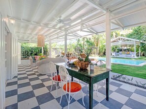 Un patio couvert doté d’un sol à carreaux, d’un espace salon extérieur, d’une piscine et d’une végétation luxuriante.