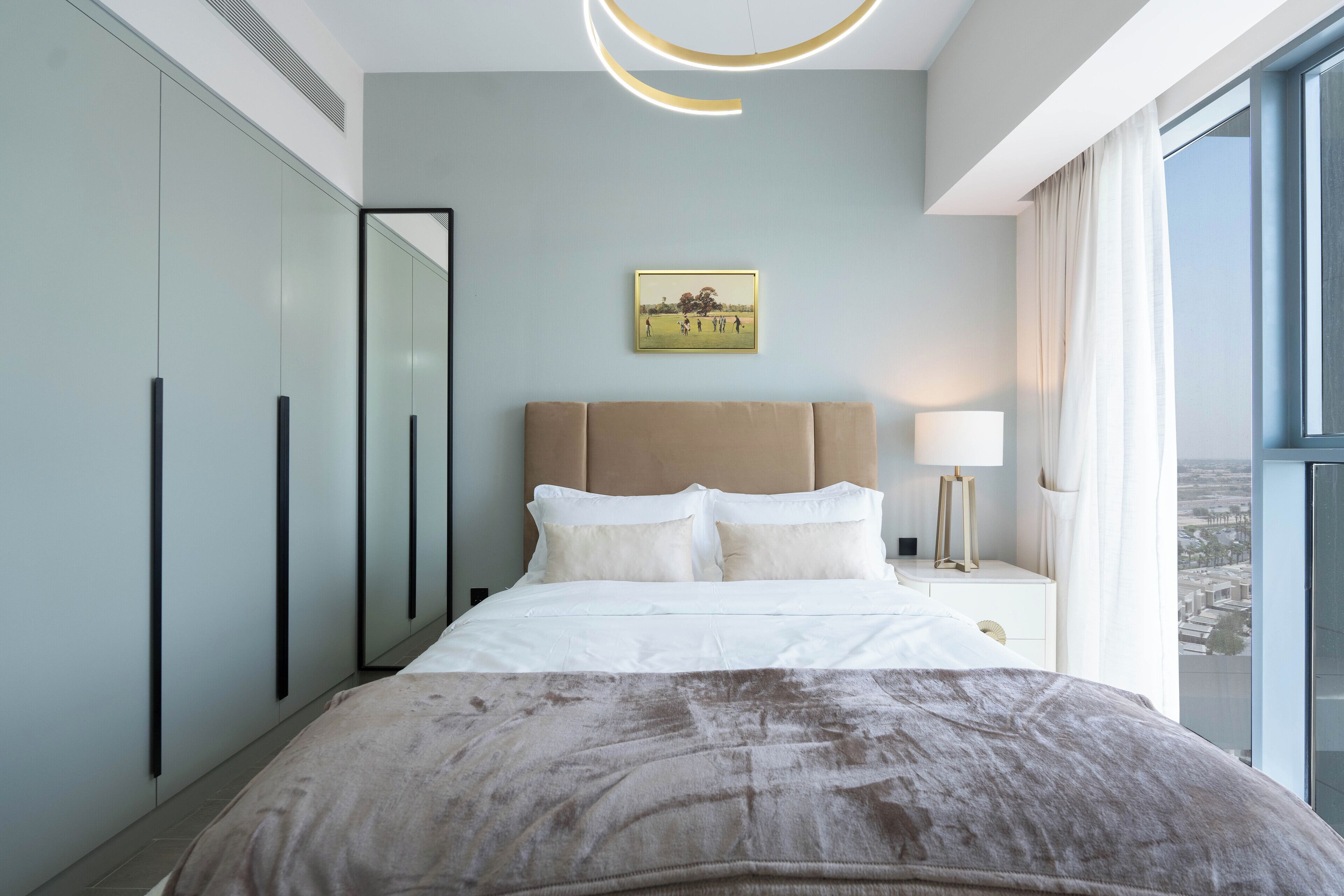 A modern hotel room with a large bed, a headboard, a bedside table with a lamp, a framed picture on the wall, and a view of the city through the window.