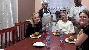 A group of people seated around a wooden dining table with plates of food, a water bottle, and a small box on the table. - Victorious Chillzone Hostel (Arusha)