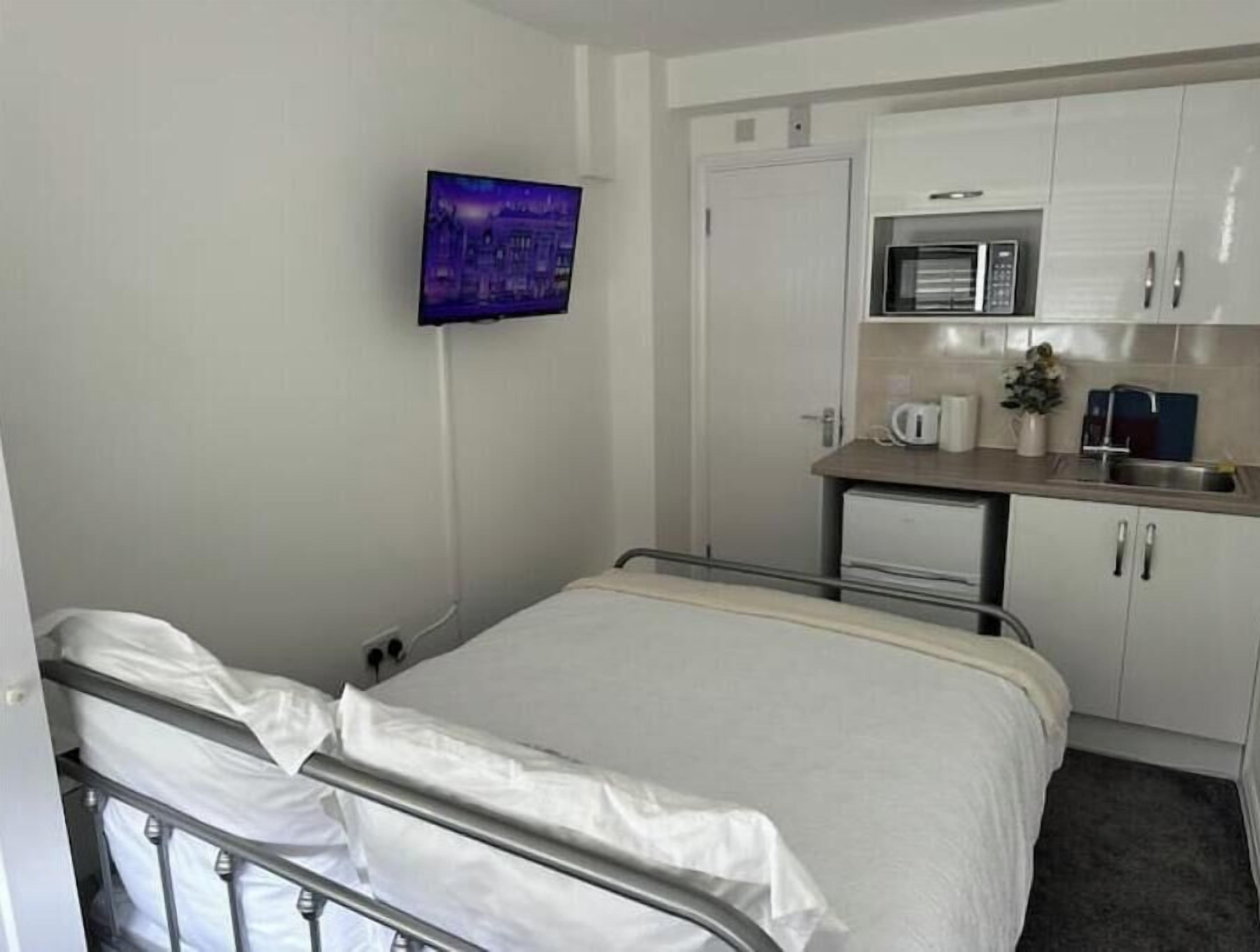A hotel room with a bed, a television mounted on the wall, a kitchenette with a microwave, and a small table with a coffee maker.