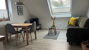 Un salón moderno con una mesa de comedor de madera, sillas grises, un sofá negro con cojines amarillos, una mesita auxiliar con un jarrón y un televisor sobre un mueble.