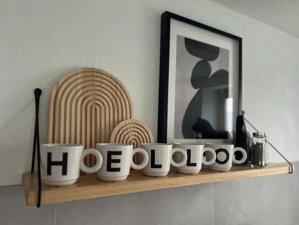 A shelf with white mugs with black letters spelling "HOELOOOO" and a framed abstract art piece on the wall.