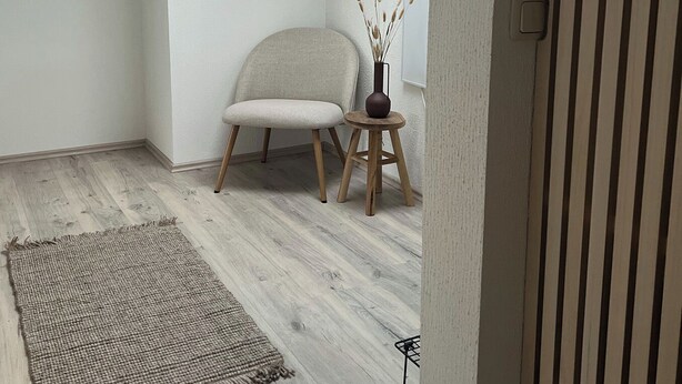 Una habitación con tragaluces, una silla, una mesita con un florero y una alfombra sobre piso de madera.