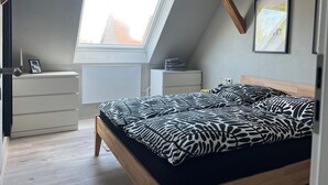 Un dormitorio con techo inclinado, tragaluz, cama con ropa de cama estampada, mesita de noche, cómoda y un cuadro en la pared.