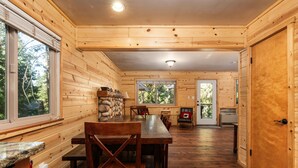 Interior de una cabaña de madera con comedor, pared de piedra y vista por una ventana.