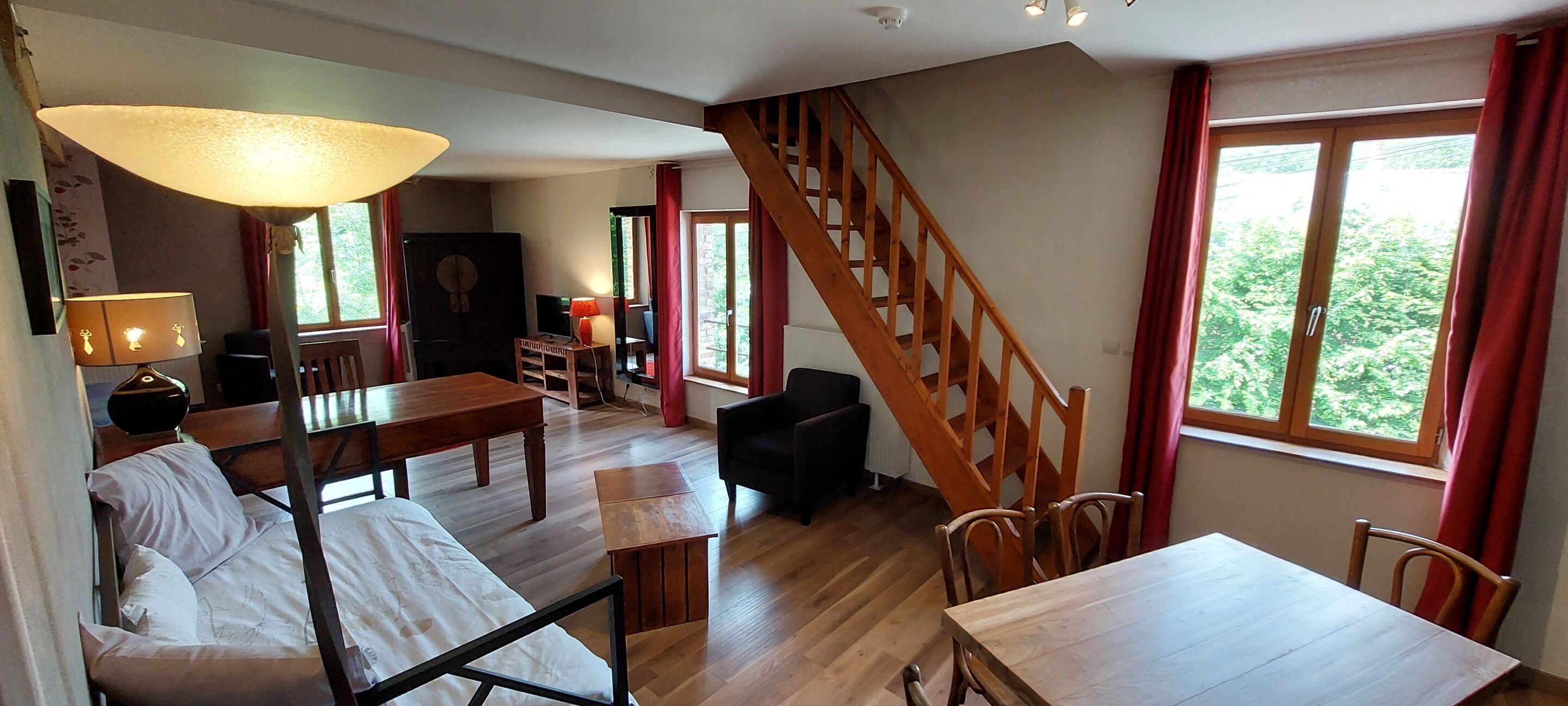 Een ruime kamer met een houten trap, een bed, een eettafel en een groot raam met rode gordijnen.