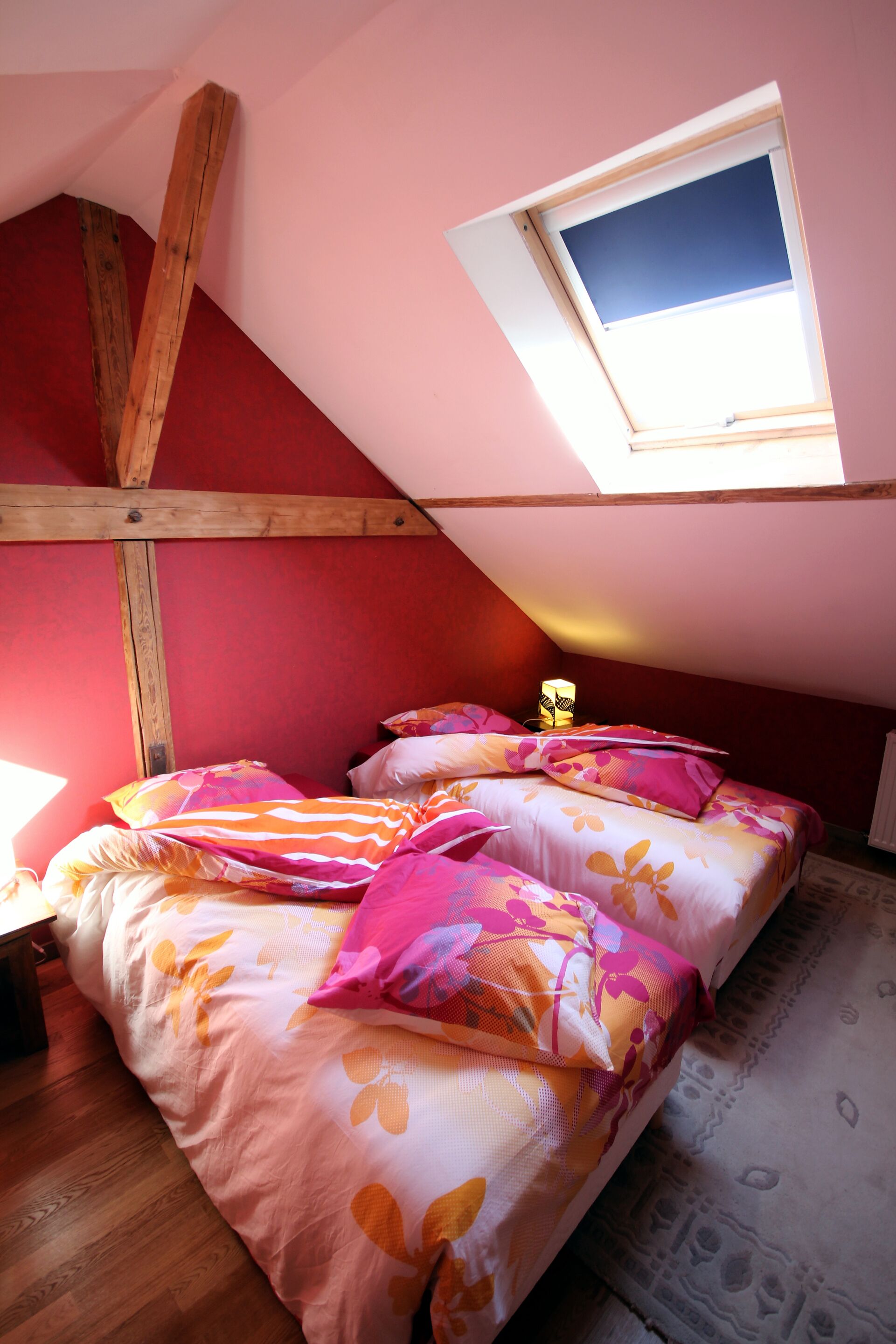 Un dormitorio con techo inclinado, tragaluz, cama con ropa de cama floral y estructura de vigas de madera.