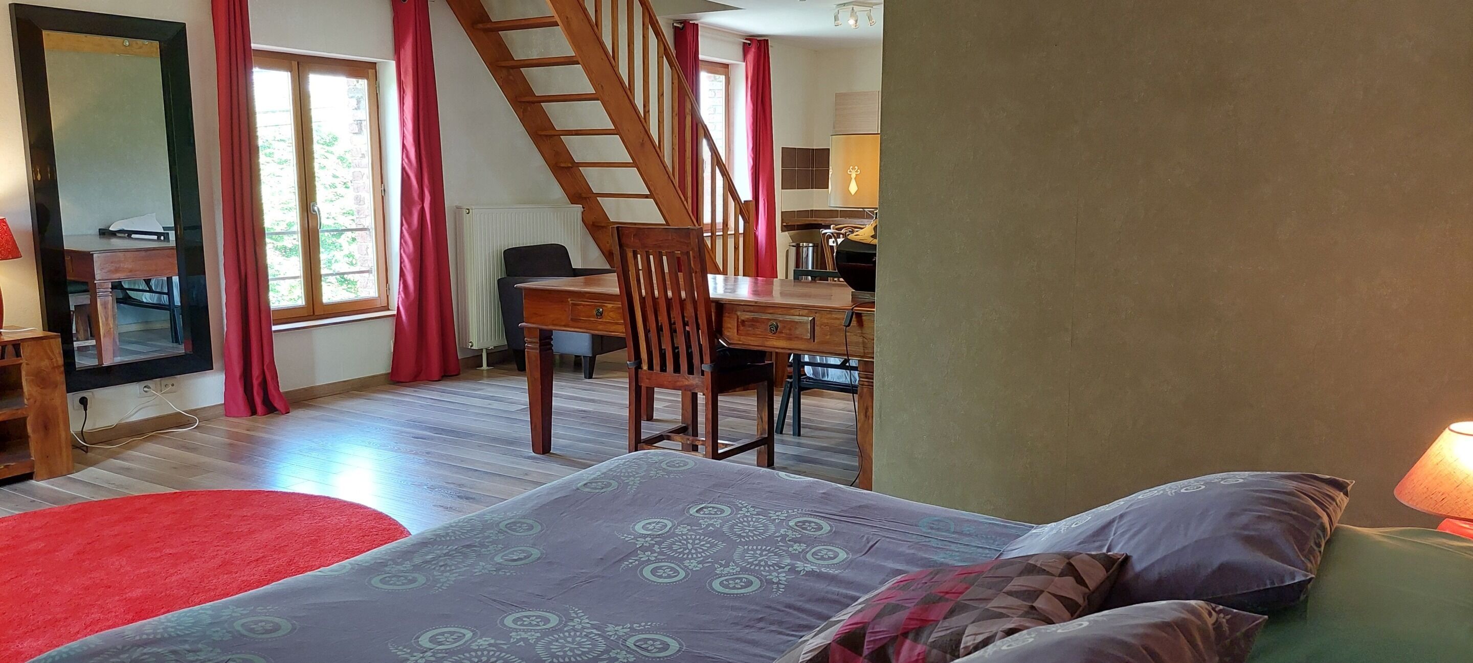 Une chambre à coucher avec un lit, un bureau et une chaise. Il y a une fenêtre avec des rideaux, un radiateur et un escalier.